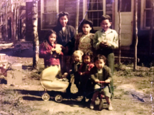 A group of seven children smiling at the camera. One is holding onto a baby doll carriage and one is on a tricycle.